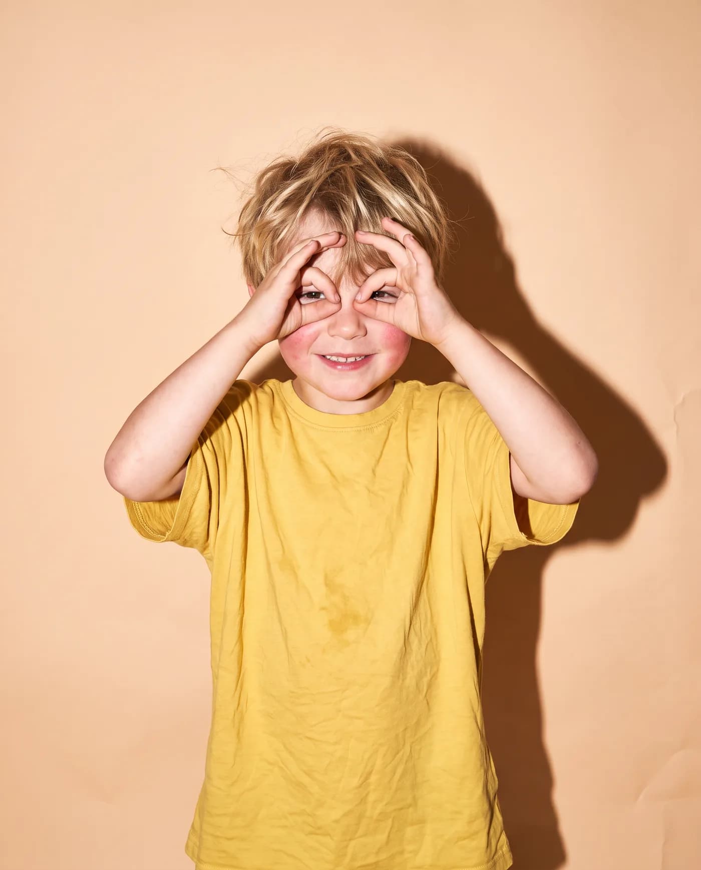 A boy on a peach backdrop makes goggles with his hands
