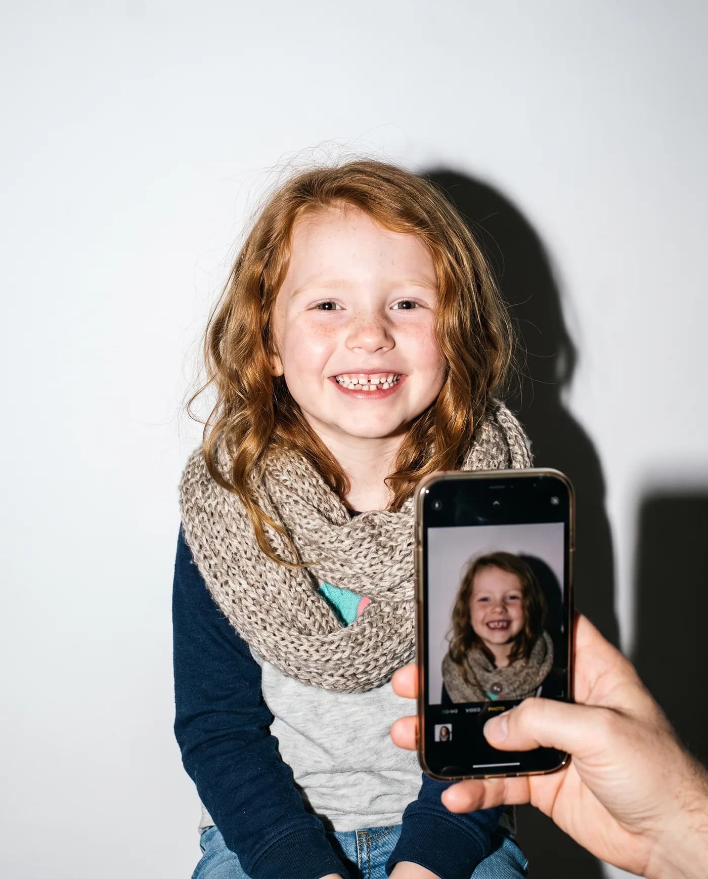 A parent’s phone photographs a grinning girl in a knit scarf on a studio backdrop