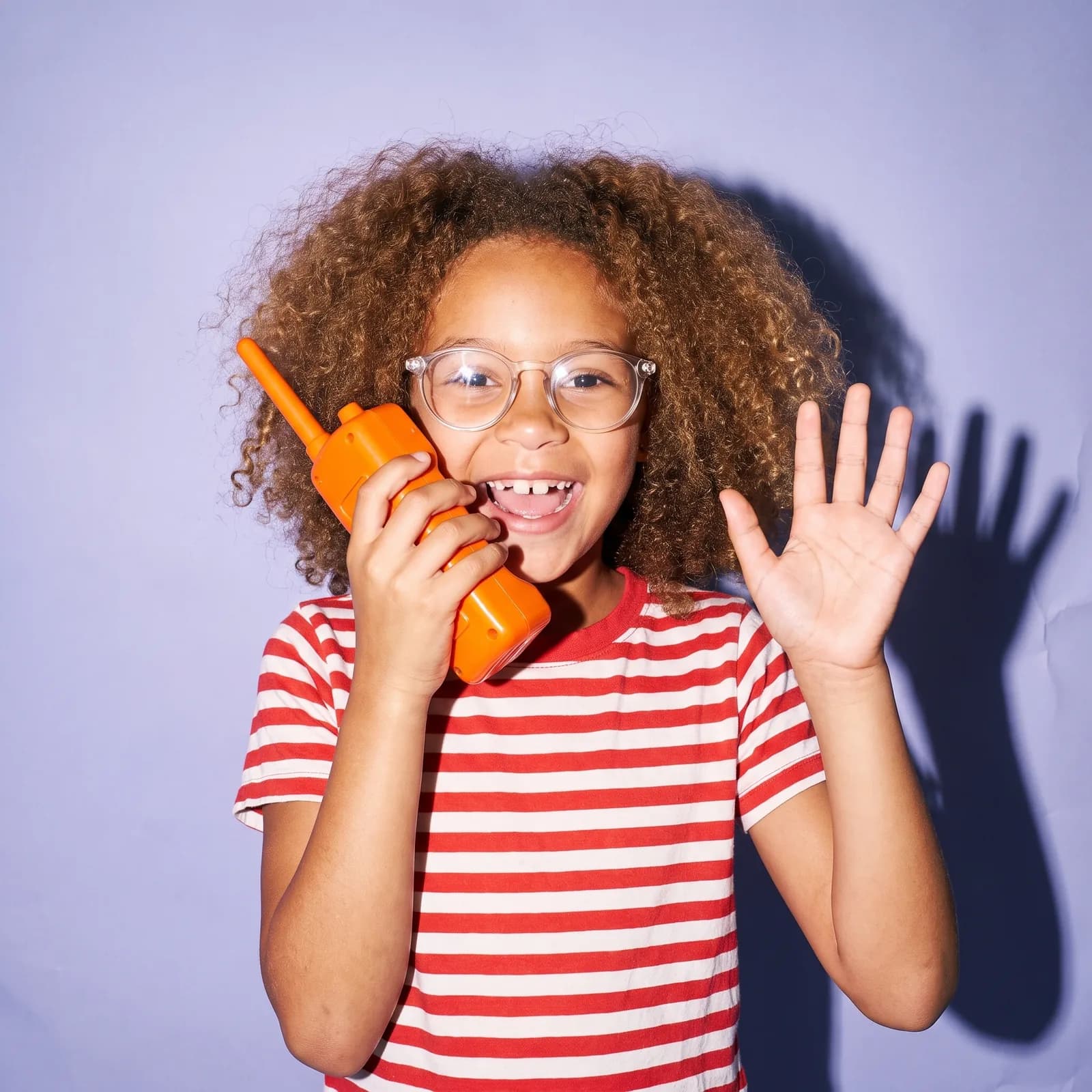 A girl on a lavender backdrop waves while talking into a toy walkie-talkie